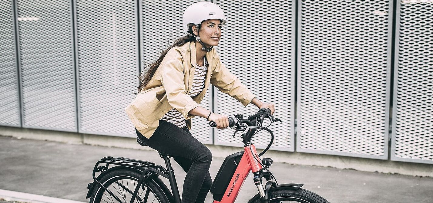 Eine Frau fährt auf einem roten Elektrorad auf Asphalt, im Hintergrund graue, gelochte Metallwände. 