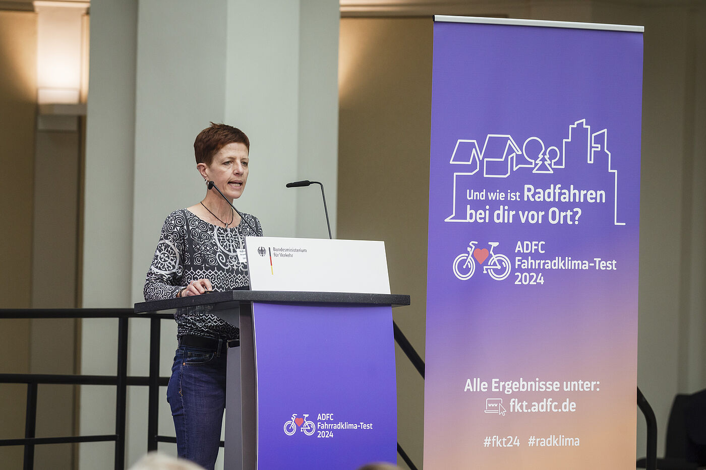 Prof. Dr. Katharina Manderscheid, Frau mit kurzen braunen Haaren in gemustertem Oberteil, steht am Rednerpult mit Mikrofon vor lila ADFC-Banner mit Aufschrift „Und wie ist Radfahren bei dir vor Ort? ADFC Fahrradklima-Test 2024"